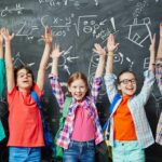 Bürgergeld: Schulbedarf und Schulmaterial im Bildungspaket enthalten 4 Kinder heben die Arme vor der Schultafel