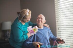 Frau umarmt pflegebedürftigen Mann im Rollstuhl mit Euro Scheinen in der Hand.