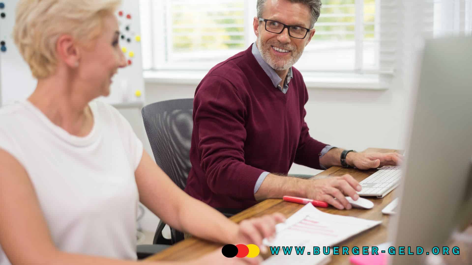 Zwei Personen sitzen gemeinsam an einem Schreibtisch in einem modernen Büro. Eine Person trägt ein weißes T-Shirt, die andere einen bordeauxroten Pullover. Beide arbeiten konzentriert am Computer, auf dem Tisch liegen Dokumente und ein Stift. Im Hintergrund sind helle Fenster und ein Whiteboard mit bunten Magneten zu sehen. Am unteren Bildrand steht die Webadresse „www.buerger-geld.org“.