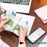 Steuererklärung für Rentner: 7 Dinge, die Sie absetzen können 5 Frau zählt 100-Euro-Scheine am Schreibtisch, vor ihr liegen Laptop, Smartphone mit Taschenrechner, offene Notizbücher und Steuerunterlagen. Auf dem Bild ist die URL www.buerger-geld.org sichtbar.