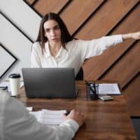 Anwälte kehren Bürgergeld-Fällen den Rücken: Was Betroffene jetzt wissen müssen 4 Anwältin im Büro zeigt mit ausgestrecktem Arm zur Seite, während sie hinter einem Laptop an einem Schreibtisch sitzt. Ihr Gegenüber sitzt ebenfalls am Tisch, Dokumente und eine Kaffeetasse liegen bereit.