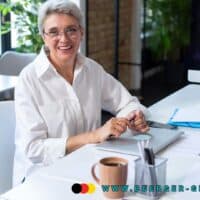 Zurück in den Job: Bundesregierung erleichtert befristete Rückkehr älterer Arbeitnehmer ab 2026 4 Ältere Frau im weißen Hemd sitzt an einem Schreibtisch im modernen Büro, vor ihr liegen Unterlagen, ein Laptop und eine Tasse Kaffee. Im Hintergrund ist ein Computerbildschirm mit Diagrammen und Text zu sehen.