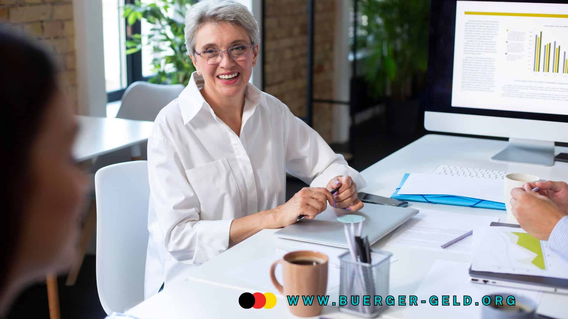 Ältere Frau im weißen Hemd sitzt an einem Schreibtisch im modernen Büro, vor ihr liegen Unterlagen, ein Laptop und eine Tasse Kaffee. Im Hintergrund ist ein Computerbildschirm mit Diagrammen und Text zu sehen.