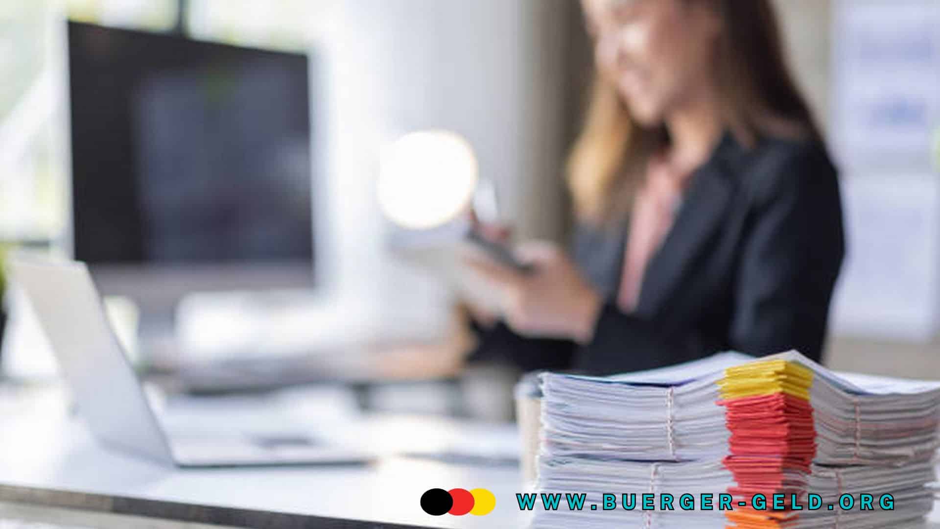 Stapel von farbig markierten Akten auf einem modernen Schreibtisch im Büro, im Hintergrund eine Person in Businesskleidung an einem Computerarbeitsplatz. Das Bild symbolisiert Büroarbeit, Verwaltung und Organisation im Kontext der Bürgergeld-Reform.