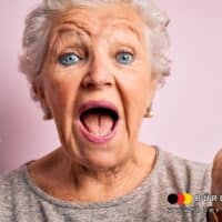 EU-Debatte: Müssen Senioren bald zum Arzt für den Führerschein? 5 Eine ältere Person mit grauen Haaren hält in ihrer linken Hand ein kleines pinkes Spielzeugauto und hebt die rechte Hand fragend oder erklärend. Die Person trägt ein hellgraues Oberteil und steht vor einem hellrosa Hintergrund. In der rechten unteren Ecke befindet sich das Logo und die Internetadresse von „Bürger & Geld“.