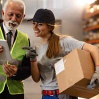 Unbegrenzter Hinzuverdienst bei Altersrente – Diese 5 Fallen lauern 5 Älterer Mann im Anzug mit Warnweste und junge Lagerarbeiterin im Gespräch beim Nebenjob in einer Lagerhalle – beide diskutieren über Arbeitsaufgaben, im Hintergrund Regale mit Kartons, Symbol für Hinzuverdienst im Ruhestand.