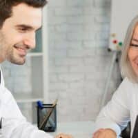 Wer eine Schwerbehinderung (GdB 50+) hat, ist oft erwerbsgemindert: Steigt jetzt auch die Erwerbsminderungsrente? 4 Ältere Frau im Gespräch mit einem Arzt am Schreibtisch, gemeinsam Betrachtung von Unterlagen zu Sozialleistungen oder Rentenanspruch.