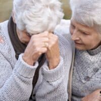 Unerwartete Krankenkassen-Falle: Wie eine kleine Auslandsrente die Familienversicherung sprengt. 5 Zwei ältere Frauen in warmer Kleidung sitzen im Freien; eine Frau wirkt verzweifelt und stützt den Kopf in die Hände, während die andere sie tröstend am Arm hält.