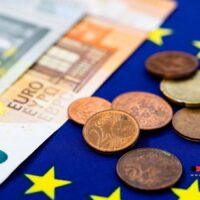 Brüssel dreht am Geldhahn – Müssen Millionen Deutsche um ihre Rente bangen? 5 Euro-Geldscheine und Münzen auf EU-Flagge – Symbolbild für geplante EU-Strategie, finanzielle Hilfen an Rentenreformen in Deutschland und Europa zu koppeln
