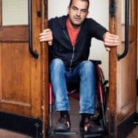 Merkzeichen G auch ohne „klassische“ Gehbehinderung: Diese Urteile ändern alles. 5 Mann im Rollstuhl öffnet schwere Holztüren in einem öffentlichen Gebäude – Symbolbild für Barrierefreiheit und den Anspruch auf Merkzeichen G bei untypischen Erkrankungen