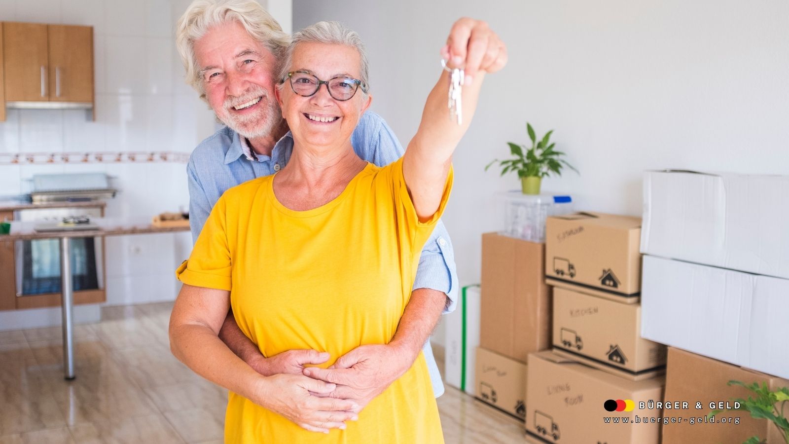Älteres Paar umarmt sich im neuen Zuhause mit Umzugskartons und hält symbolisch einen Wohnungsschlüssel – Rentner mit Wohnproblemen und Wohngeld-Anspruch.