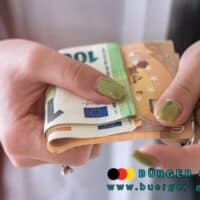 Nahaufnahme von Händen mit grün lackierten Fingernägeln, die mehrere Euro-Geldscheine halten; unten im Bild das Logo und die Webadresse von BÜRGER & GELD (www.buerger-geld.org).