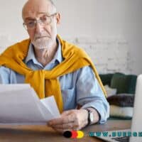 Älterer Mann sitzt am Tisch in einem Wohnzimmer, trägt ein hellblaues Hemd und einen gelben Pullover über den Schultern. Er hält mehrere Dokumente in der Hand und schaut auf Papiere, vor ihm steht ein Laptop. Im Hintergrund ist ein Sofa und eine helle Wand zu sehen. Unten rechts befindet sich das Logo von buerger-geld.org.