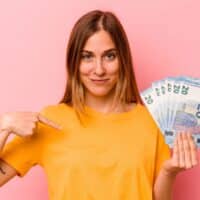 unge Frau im gelben T-Shirt hält lächelnd mehrere 20-Euro-Scheine in der Hand und zeigt mit dem Finger auf sich vor rosa Hintergrund.