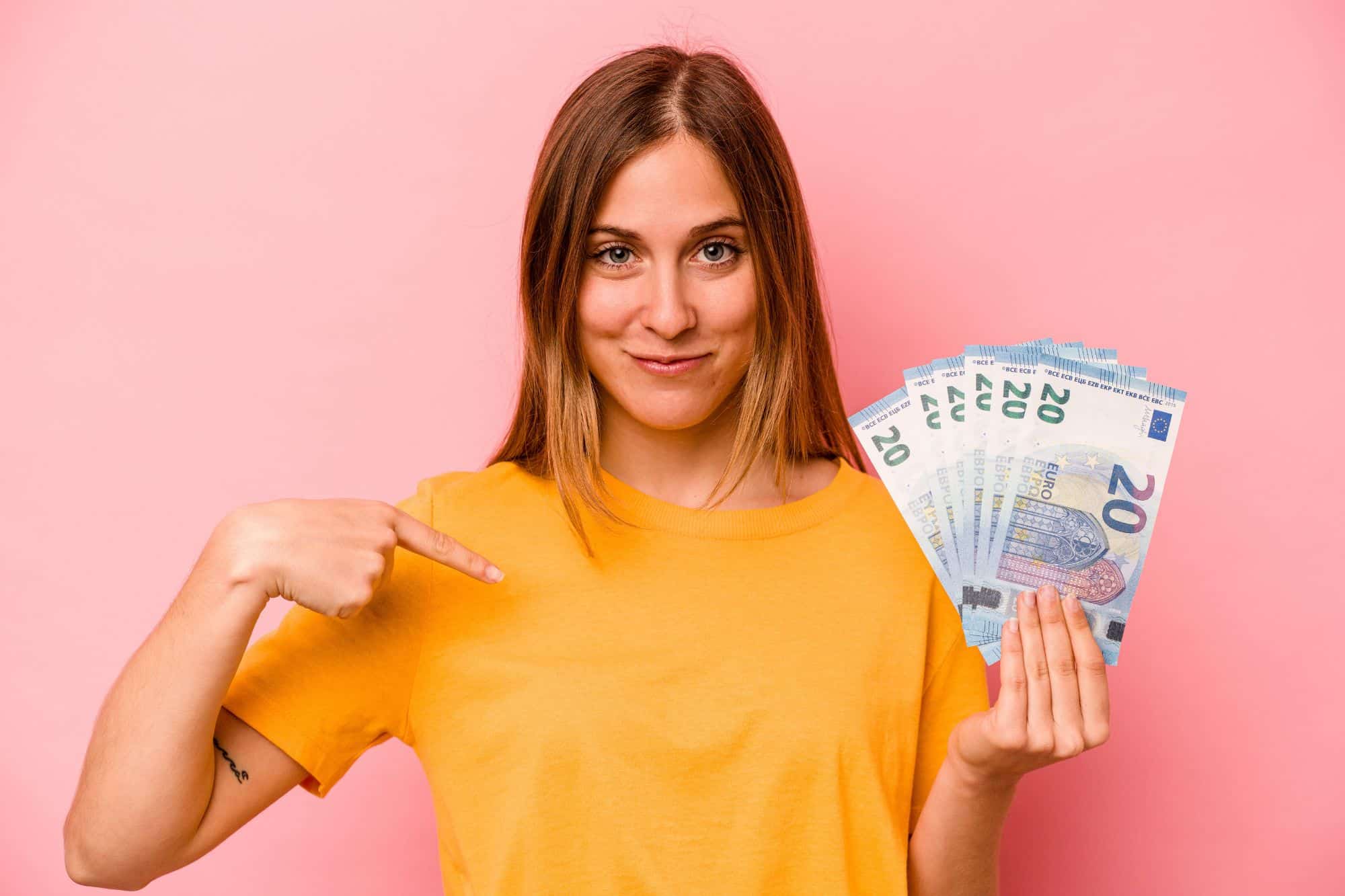 unge Frau im gelben T-Shirt hält lächelnd mehrere 20-Euro-Scheine in der Hand und zeigt mit dem Finger auf sich vor rosa Hintergrund.