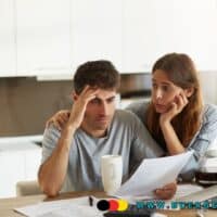 Ein junges Paar sitzt gemeinsam am Küchentisch und prüft Unterlagen zu Finanzen und Kontopfändung. Auf dem Tisch liegen Dokumente, ein Laptop, eine Kaffeekanne und eine Tasse. Im Vordergrund ist das Logo von Bürger-Geld.org zu sehen.