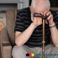 Älterer Mann mit weißem Haar sitzt nachdenklich auf einem Sessel und stützt sich mit beiden Händen auf einen Gehstock. Im Hintergrund ist ein Kamin zu sehen. Die Szene wirkt ruhig, aber nachdenklich und symbolisiert Unsicherheit im Alter.