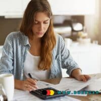 Frau sitzt am Küchentisch, prüft Finanzunterlagen und tippt auf einem Taschenrechner neben Laptop und Kaffeetasse, Symbolbild für Haushaltsfinanzen und Umschuldung, im Vordergrund das Logo von buerger-geld.org.