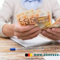 Eine ältere Person zählt mehrere 50-Euro-Geldscheine am Schreibtisch, daneben liegen ein Taschenrechner, ein Kugelschreiber und ein Notizblock. Unten im Bild steht die Webadresse www.buerger-geld.org.