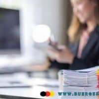 Stapel von farbig markierten Akten auf einem modernen Schreibtisch im Büro, im Hintergrund eine Person in Businesskleidung an einem Computerarbeitsplatz. Das Bild symbolisiert Büroarbeit, Verwaltung und Organisation im Kontext der Bürgergeld-Reform.