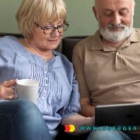 Seniorenpaar sitzt gemeinsam auf dem Sofa, schaut entspannt auf ein Tablet und trinkt Kaffee; eingeblendete Webadresse www.buerger-geld.org.