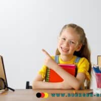 Mädchen mit Zöpfen im gelben Shirt sitzt an einem Schreibtisch, hält ein rotes Notizbuch im Arm und zeigt mit dem Finger nach links; auf dem Tisch liegen Bücher, eine Schuluhr, ein Apfel, Stifte sowie eine blaue Schultasche.