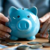 Blaues Sparschwein wird von zwei Händen gehalten, umgeben von Münzen und EC-Karten – symbolisch für private Altersvorsorge und finanziellen Aufbau.