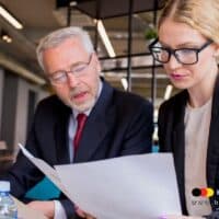 Zwei Personen im Business-Outfit prüfen gemeinsam wichtige Dokumente zur Rentenbesteuerung, im modernen Büro mit Logo von Bürger & Geld, www.buerger-geld.org.