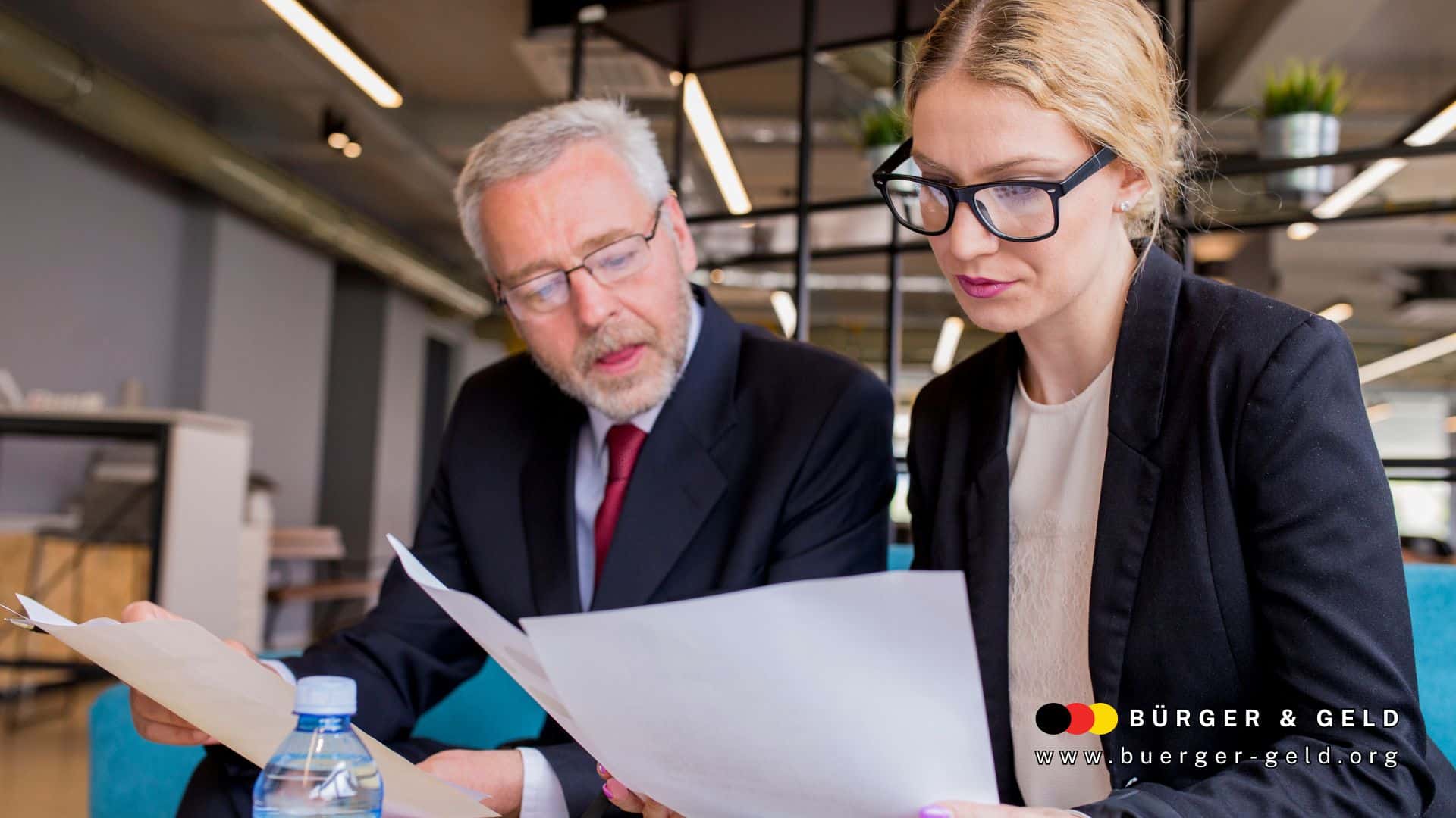 Zwei Personen im Business-Outfit prüfen gemeinsam wichtige Dokumente zur Rentenbesteuerung, im modernen Büro mit Logo von Bürger & Geld, www.buerger-geld.org.