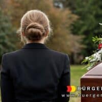 Eine Frau in schwarzer Trauerkleidung steht bei einer Beerdigung vor einem geschlossenen Sarg mit Blumenarrangement. Im Hintergrund ist ein weißes Holzkreuz zu sehen. Im Bild unten rechts befindet sich das Logo von BÜRGER & GELD sowie die Website-Adresse www.buerger-geld.org.