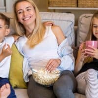 Mutter sitzt mit zwei lachenden Grundschulkindern auf dem Sofa mit einer Scüssel Popcorn, im Hintergrund ein großes transparentes Euro-Zeichen.