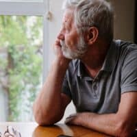 Älterer Mann sitzt nachdenklich an einem Tisch vor einem Fenster, auf dem Tisch liegen eine Brille und ein Smartphone.
