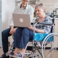 Seniorin im Rollstuhl mit Laptop, unterstützt von einem Mann, in einer modernen Küche; im Vordergrund Scheine Euro-Geld, Symbol für finanzielle Zuschüsse bei Grundsicherung, Pflegegrad und Schwerbehinderung.