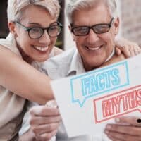 Älteres, glückliches Paar mit Brille sitzt zusammen und hält ein Papier mit der Aufschrift „Facts“ und „Myths“ in den Händen – Symbolbild für Faktencheck bei Rentenmythen, Bürger & Geld Magazin.