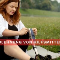Eine Frau mit einer Beinprothese sitzt auf einer Wiese und passt das Hilfsmittel an. Im Vordergrund ist der Schriftzug „Ablehnung von Hilfsmitteln“ zu sehen. Logo und Webadresse von Bürger & Geld erscheinen unten rechts.