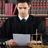 Richter im Talar sitzt mit gerichtlichem Urteil und Gesetzbüchern im Hintergrund, vor ihm liegen Richterhammer und Waage, Symbol der Justiz, sowie das Bürger & Geld Logo.
