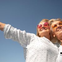 Zwei Seniorinnen mit Sonnenbrillen lächeln glücklich in den blauen Himmel – Symbol für frühere Rentenzahlung und neue Verbraucherregeln ab November 2025.