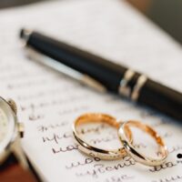 Zwei Eheringe, ein Füller und eine Uhr liegen auf einem handgeschriebenen Brief – Symbol für Witwerrente, Ehe und Rechtsentscheidung.