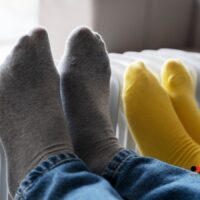 Zwei Personen wärmen ihre Füße in Socken an einer weißen Heizung – Symbolbild für Heizkosten und Betriebsstrom der Gastherme beim Bürgergeld.