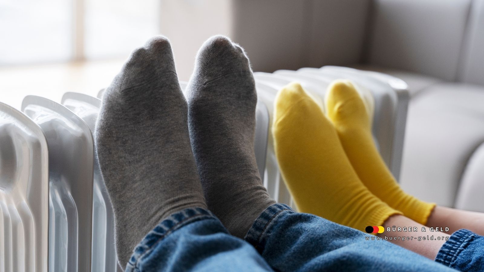 Zwei Personen wärmen ihre Füße in Socken an einer weißen Heizung – Symbolbild für Heizkosten und Betriebsstrom der Gastherme beim Bürgergeld.