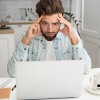 Ein sichtlich besorgter Mann sitzt am Küchentisch vor seinem Laptop und hält sich den Kopf, während er einen Brief von der Krankenkasse liest. Auf dem Tisch liegen eine Kaffeetasse, ein Notizbuch und ein Kugelschreiber.