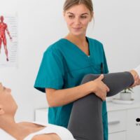 Physiotherapeutin unterstützt Patientin bei Reha-Übung im Behandlungsraum, im Hintergrund sind anatomische Poster zu sehen.