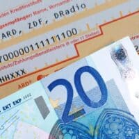 Überweisungsformular für den Rundfunkbeitrag (ARD, ZDF, Deutschlandradio) mit sichtbar ausgefüllter IBAN und einem zwanzig Euro-Schein davor als Symbol für die Zahlungsweise.