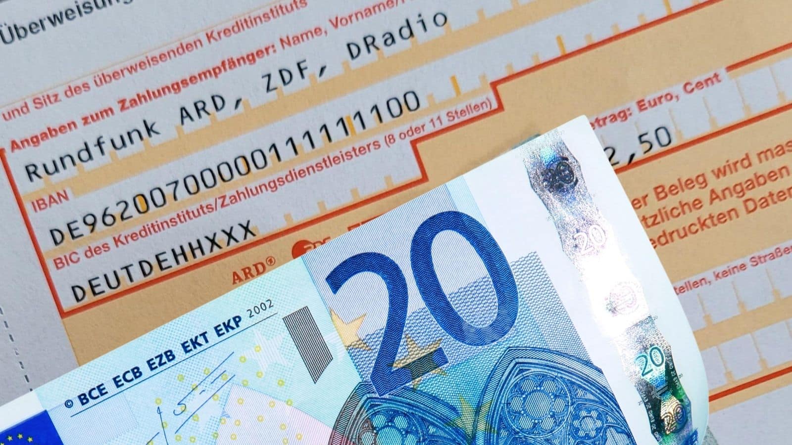 Überweisungsformular für den Rundfunkbeitrag (ARD, ZDF, Deutschlandradio) mit sichtbar ausgefüllter IBAN und einem zwanzig Euro-Schein davor als Symbol für die Zahlungsweise.