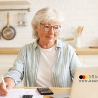 Ältere Frau sitzt am Küchentisch und prüft Unterlagen vor Laptop, Taschenrechner und Kaffeetasse – Finanzplanung und Online-Anwendungen im Ruhestand