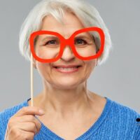 Ältere schwerbehinderte Frau mit weißem Haar hält eine rote Brillenattrappe vor ihr Gesicht, Symbol für das Thema Brillenkosten und fehlende Kostenübernahme durch Krankenkasse oder Stadt in Deutschland.