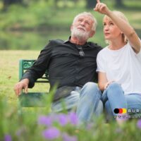 Älteres Paar sitzt entspannt auf einer grünen Parkbank im Freien und genießt den Ruhestand. Die Frau zeigt auf etwas in der Ferne, beide wirken gelöst und glücklich. Im Vordergrund blühen lila Blumen; unten rechts Logo und URL von BÜRGER & GELD sichtbar.