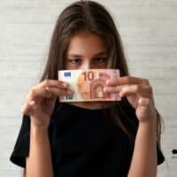 Junge Frau mit langen braunen Haaren hält einen Geldschein in beiden Händen; ihr Gesicht ist unkenntlich gemacht. Im Vordergrund steht das Thema Kindergeld; unten rechts ist das Logo des Nachrichtenportals BÜRGER & GELD zu sehen.