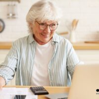 Rentnerin sitzt am Küchentisch und prüft Unterlagen zur Krankenversicherung im Ruhestand, während sie mit Taschenrechner die Kosten kalkuliert; Bürger & Geld Nachrichtenportal Logo sichtbar.