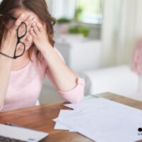 Frau am Tisch, hält sich erschöpft das Gesicht, Dokumente und Laptop vor sich – Kündigungsschutz bei Behinderung, Bürger & Geld Nachrichtenmagazin